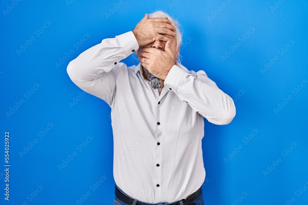 Middle age man with grey hair standing over blue background covering eyes and mouth with hands, surprised and shocked. hiding emotion