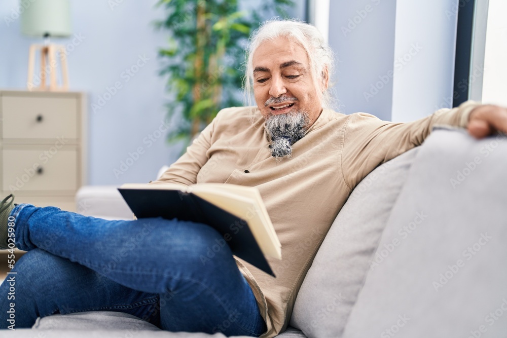 Middle age grey-haired man reading book sitting on sofa at home Middle age grey-haired man reading book sitting on sofa at home