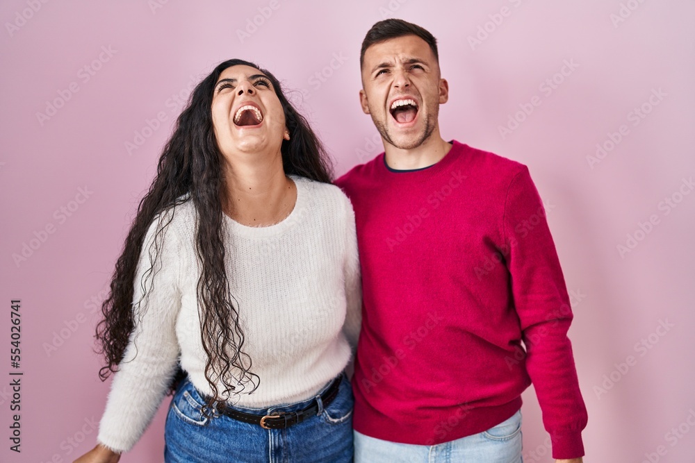 Young hispanic couple standing over pink background angry and mad screaming frustrated and furious, shouting with anger. rage and aggressive concept. Young hispanic couple standing over pink background angry and mad screaming frustrated and furious, shouting with anger. rage and aggressive concept.