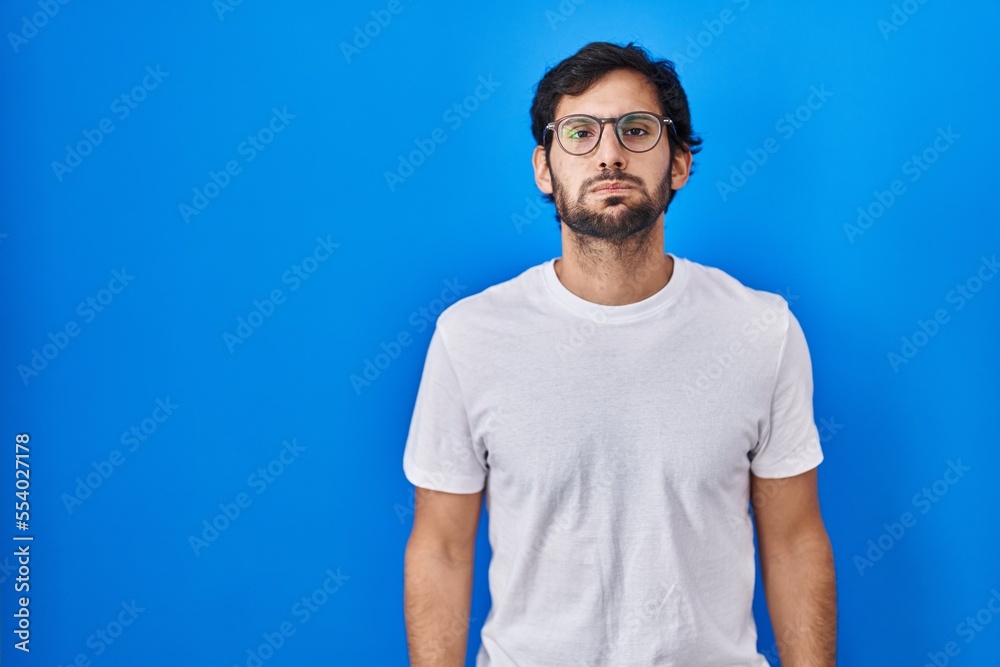 Handsome latin man standing over blue background puffing cheeks with funny face. mouth inflated with air, crazy expression. Handsome latin man standing over blue background puffing cheeks with funny face. mouth inflated with air, crazy expression.