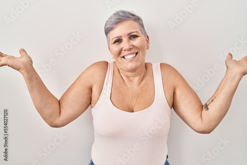 Middle age caucasian woman standing over white background clueless and confused expression with arms and hands raised. doubt concept.