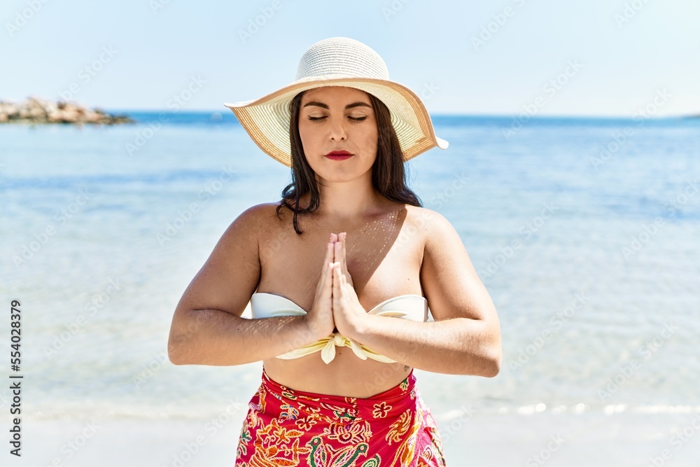 Young beautiful hispanic woman tourist wearing bikini and summer hat doing yoga exercise at seaside Young beautiful hispanic woman tourist wearing bikini and summer hat doing yoga exercise at seaside