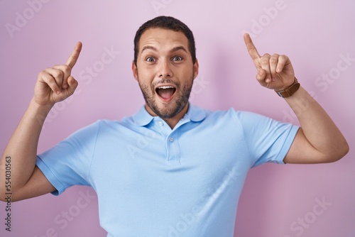 Hispanic man standing over pink background smiling amazed and surprised and pointing up with fingers and raised arms.