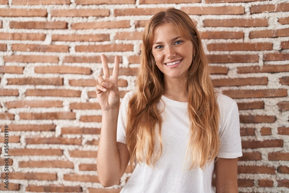 Young caucasian woman standing over bricks wall showing and pointing up with fingers number two while smiling confident and happy. Young caucasian woman standing over bricks wall showing and pointing up with fingers number two while smiling confident and happy.