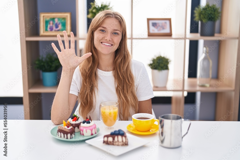 Young caucasian woman eating pastries t for breakfast showing and pointing up with fingers number five while smiling confident and happy.