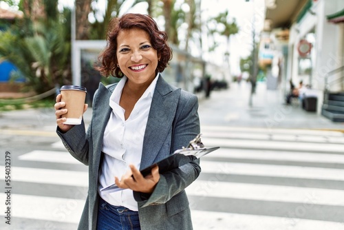 Middle age latin businesswoman drinking coffee and holding clipboard at the city.