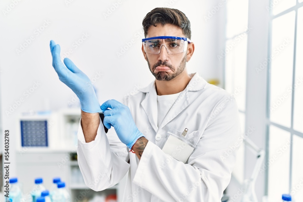 Young hispanic man working at scientist laboratory putting gloves on depressed and worry for distress, crying angry and afraid. sad expression. Young hispanic man working at scientist laboratory putting gloves on depressed and worry for distress, crying angry and afraid. sad expression.