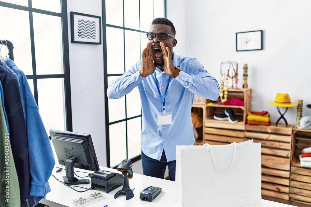 Young african man working as manager at retail boutique shouting angry out loud with hands over mouth Young african man working as manager at retail boutique shouting angry out loud with hands over mouth