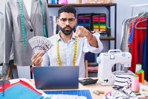 Hispanic man with beard dressmaker designer holding dollars pointing with finger to the camera and to you, confident gesture looking serious