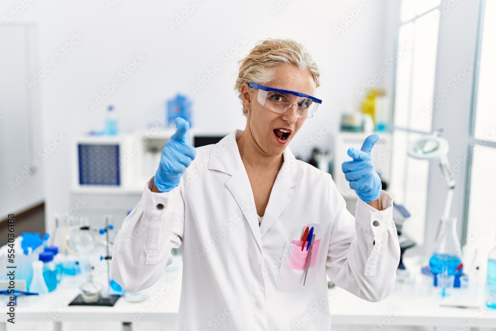 Middle age blonde woman working at scientist laboratory pointing fingers to camera with happy and funny face. good energy and vibes.