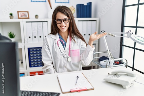 Young doctor woman wearing doctor uniform and stethoscope at the clinic smiling cheerful presenting and pointing with palm of hand looking at the camera.