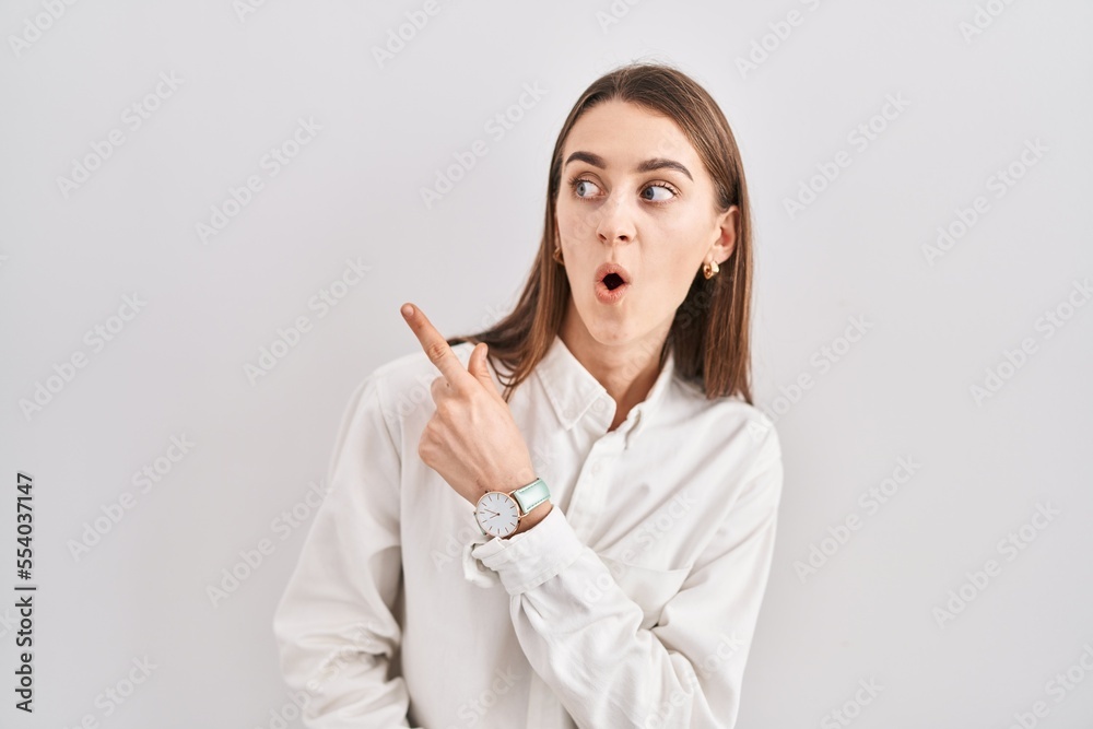 Young caucasian woman standing over isolated background surprised pointing with finger to the side, open mouth amazed expression. Young caucasian woman standing over isolated background surprised pointing with finger to the side, open mouth amazed expression.