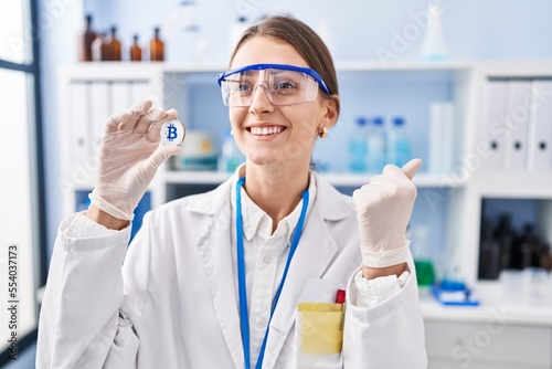 Young caucasian woman working at scientist laboratory holding bitcoin screaming proud, celebrating victory and success very excited with raised arms