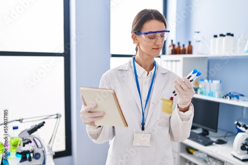 Young caucasian woman scientist holding test tubes reading notebook at laboratory