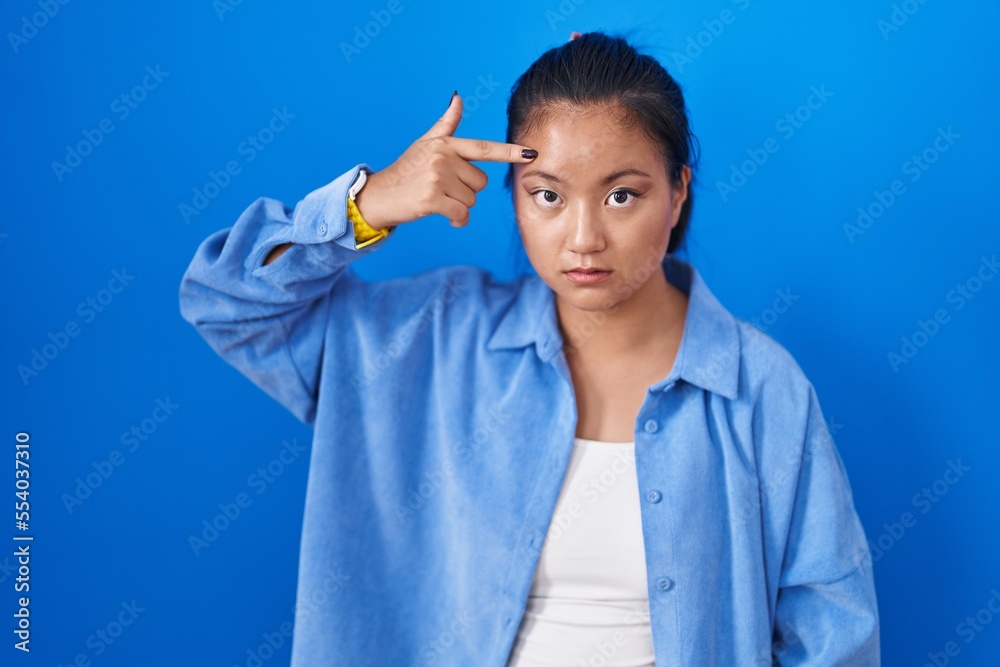 Asian young woman standing over blue background pointing unhappy to pimple on forehead, ugly infection of blackhead. acne and skin problem Asian young woman standing over blue background pointing unhappy to pimple on forehead, ugly infection of blackhead. acne and skin problem