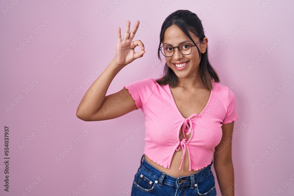 Hispanic young woman standing over pink background wearing glasses smiling positive doing ok sign with hand and fingers. successful expression. Hispanic young woman standing over pink background wearing glasses smiling positive doing ok sign with hand and fingers. successful expression.