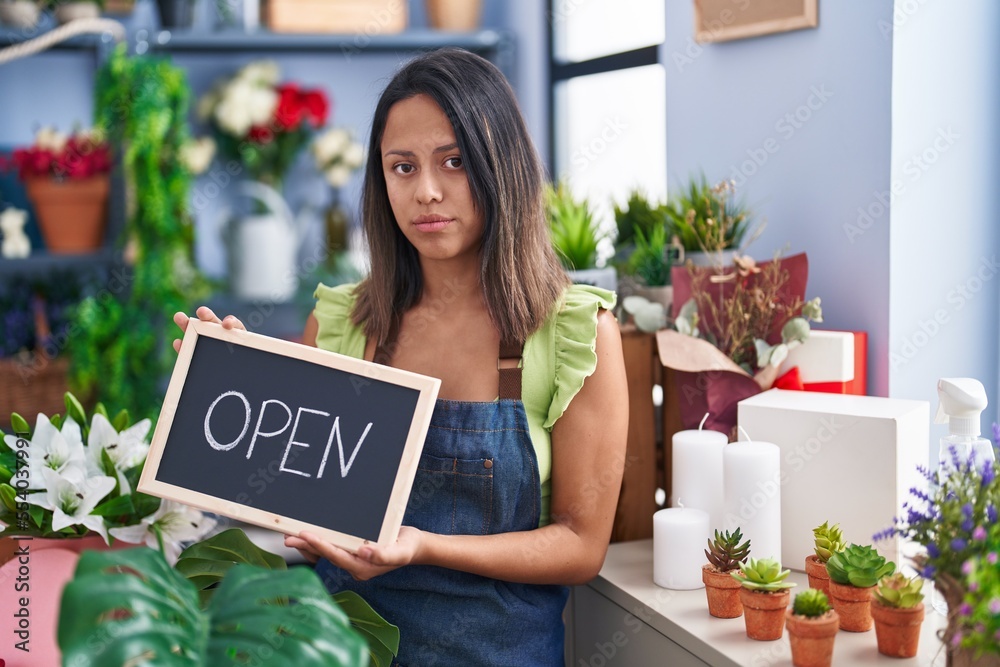 Hispanic young woman working at florist with open sign relaxed with serious expression on face. simple and natural looking at the camera. Hispanic young woman working at florist with open sign relaxed with serious expression on face. simple and natural looking at the camera.
