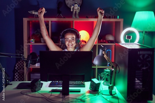 Young beautiful hispanic woman streamer playing video game with winner expression at gaming room