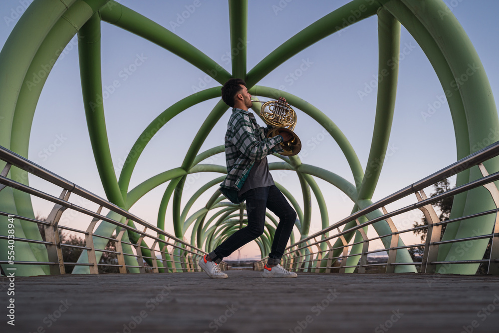 Musician walking on a bridge with his horn in his hand.