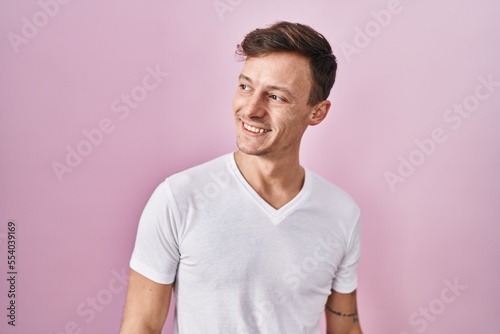 Caucasian man standing over pink background looking away to side with smile on face, natural expression. laughing confident.