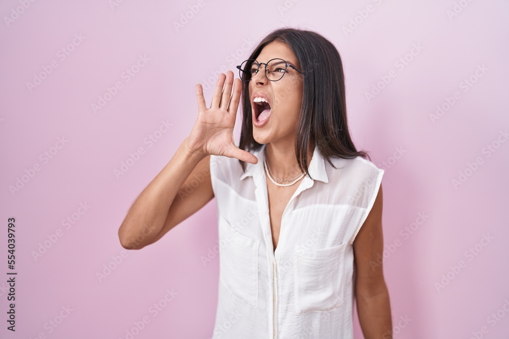 Brunette young woman standing over pink background wearing glasses shouting and screaming loud to side with hand on mouth. communication concept. Brunette young woman standing over pink background wearing glasses shouting and screaming loud to side with hand on mouth. communication concept.