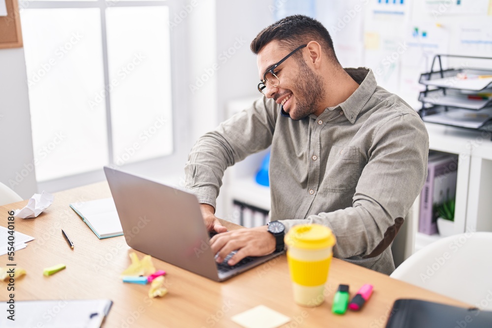 Young hispanic man business worker using laptop talking on smartphone at office Young hispanic man business worker using laptop talking on smartphone at office