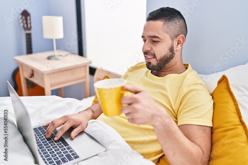 Young latin man using laptop drinking coffee at bedroom