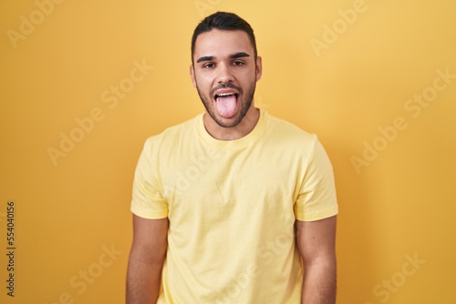 Young hispanic man standing over yellow background sticking tongue out happy with funny expression. emotion concept.