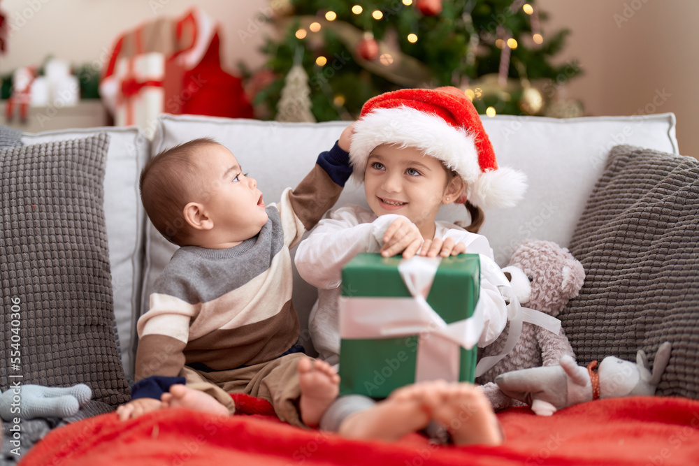 Brother and sister opening gift sitting on sofa by christmas tree at home Brother and sister opening gift sitting on sofa by christmas tree at home