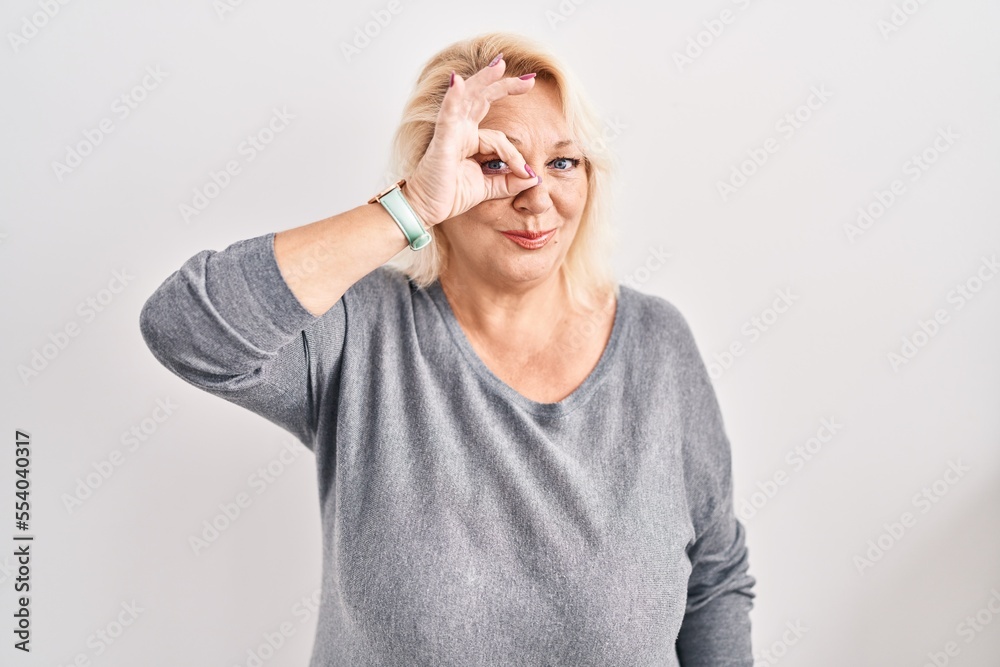 Middle age caucasian woman standing over white background doing ok gesture with hand smiling, eye looking through fingers with happy face.