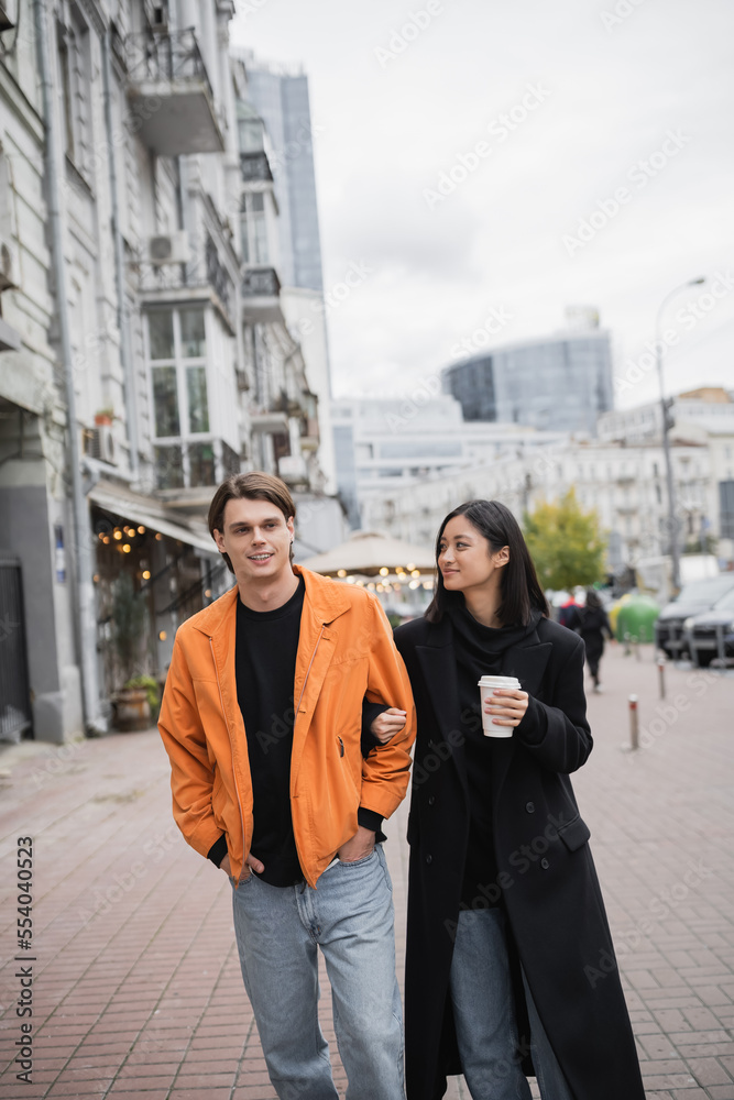 Smiling asian woman holding coffee to go and looking at boyfriend on urban street.