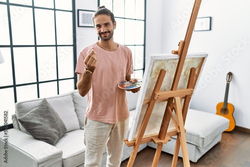 Young hispanic man with beard painting on canvas at home doing money gesture with hands, asking for salary payment, millionaire business