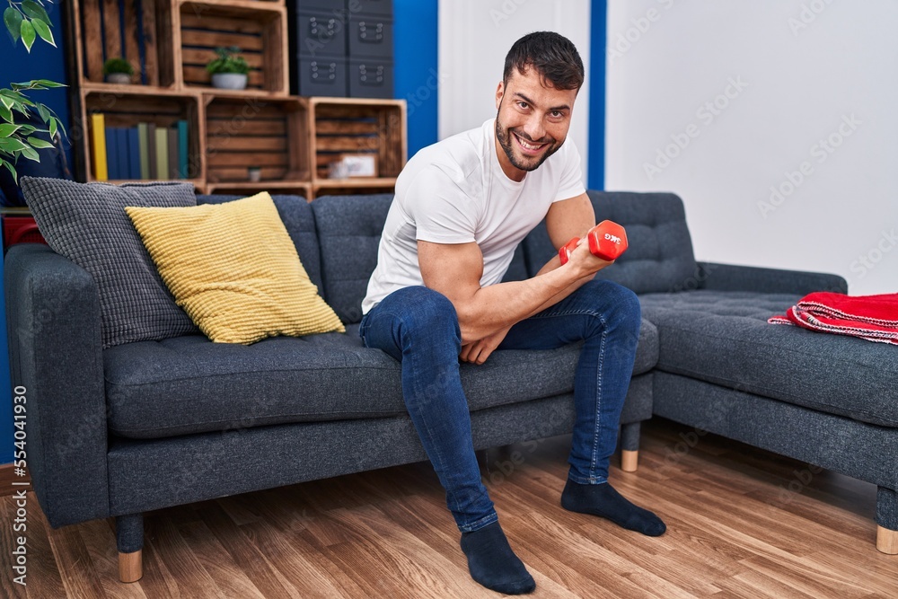 Young hispanic man training using dumbbell sitting on sofa at home