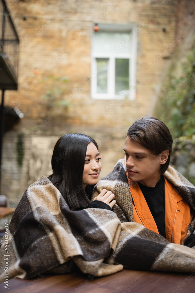 Obraz premium Young multiethnic couple in blanket looking at each other in outdoor cafe.