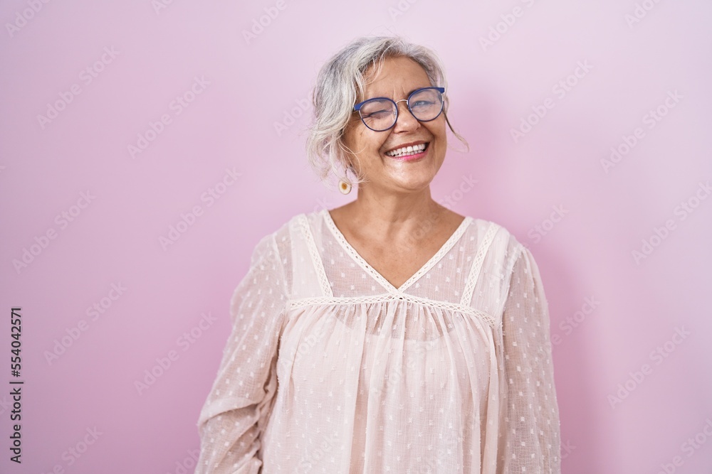 Middle age woman with grey hair standing over pink background winking looking at the camera with sexy expression, cheerful and happy face.