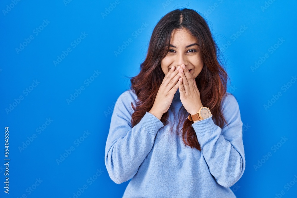Hispanic young woman standing over blue background laughing and embarrassed giggle covering mouth with hands, gossip and scandal concept Hispanic young woman standing over blue background laughing and embarrassed giggle covering mouth with hands, gossip and scandal concept