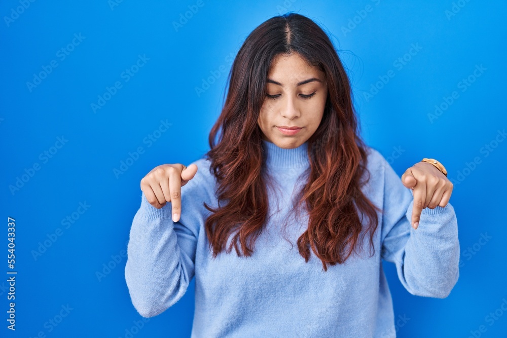 Hispanic young woman standing over blue background pointing down looking sad and upset, indicating direction with fingers, unhappy and depressed. Hispanic young woman standing over blue background pointing down looking sad and upset, indicating direction with fingers, unhappy and depressed.