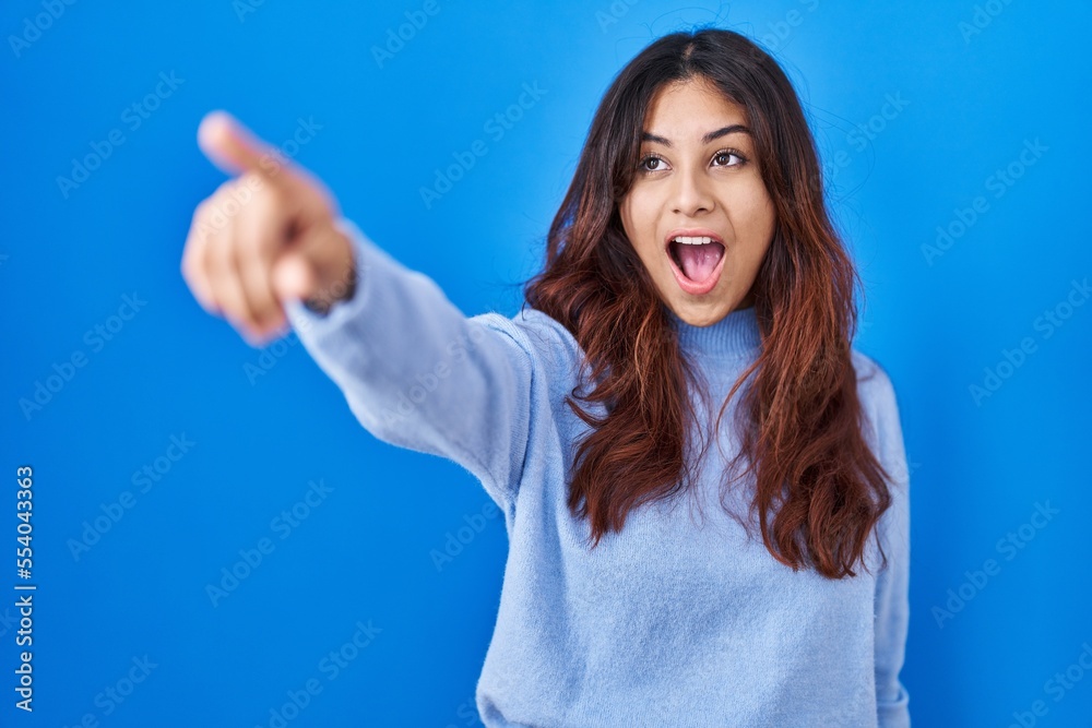 Fototapeta premium Hispanic young woman standing over blue background pointing with finger surprised ahead, open mouth amazed expression, something on the front