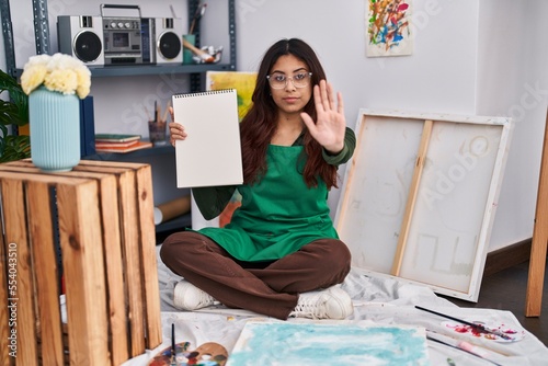 Hispanic young woman holding notebook at art studio with open hand doing stop sign with serious and confident expression, defense gesture