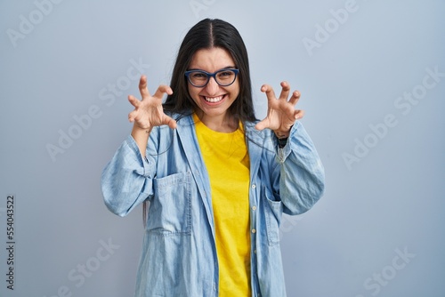 Young hispanic woman standing over blue background smiling funny doing claw gesture as cat, aggressive and sexy expression