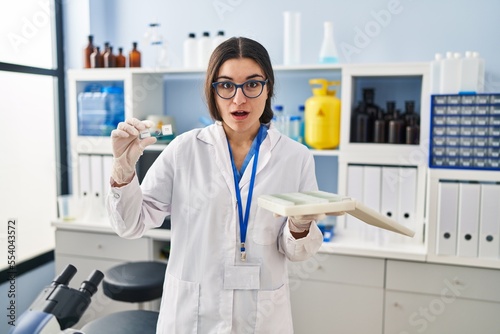 Young hispanic woman working at scientist laboratory with blood samples afraid and shocked with surprise and amazed expression  fear and excited face.