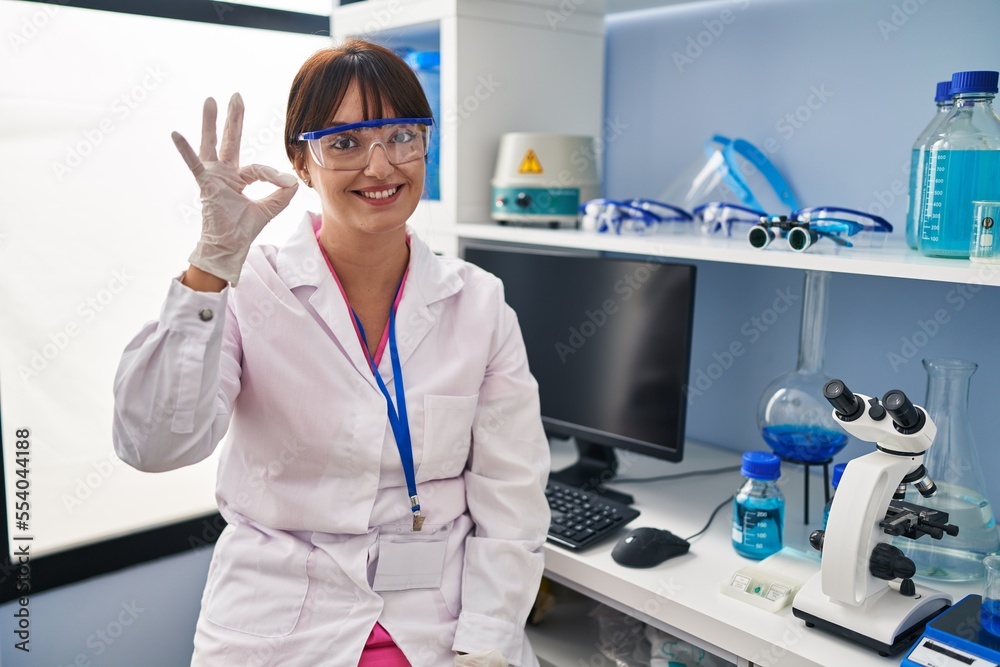 Young brunette woman working at scientist laboratory smiling positive doing ok sign with hand and fingers. successful expression. Young brunette woman working at scientist laboratory smiling positive doing ok sign with hand and fingers. successful expression.