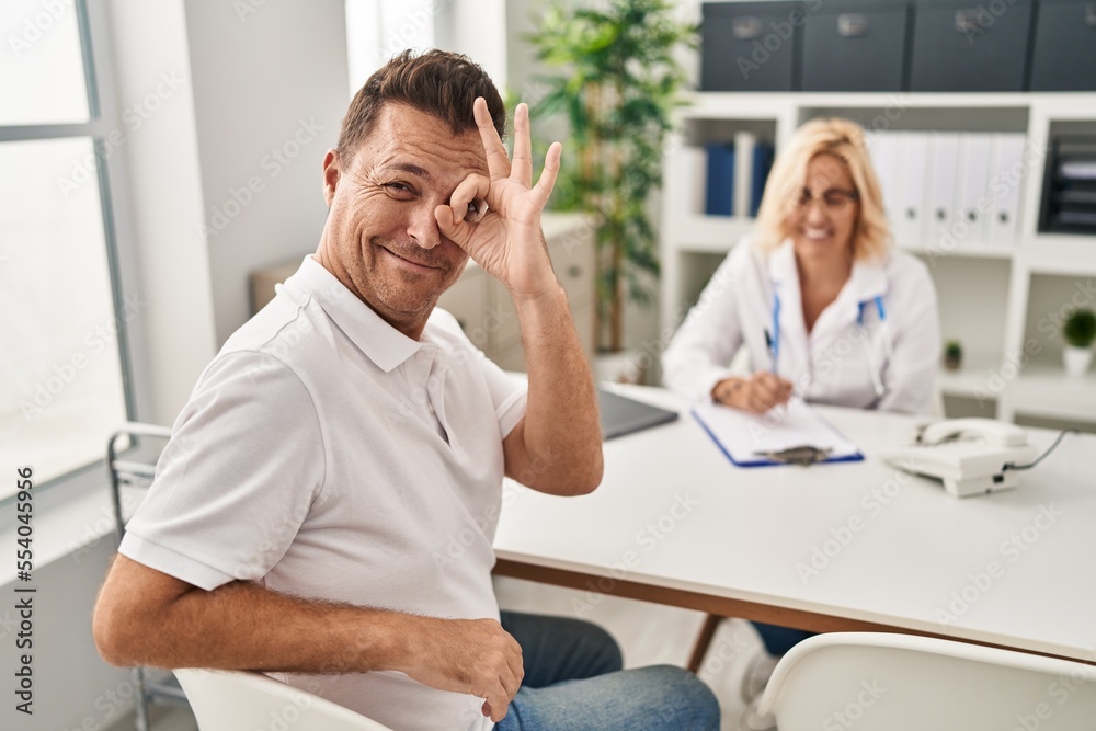 Hispanic man at the doctor smiling happy doing ok sign with hand on eye looking through fingers Hispanic man at the doctor smiling happy doing ok sign with hand on eye looking through fingers