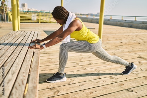 African american woman wearing sportswear stretching leg at seaside