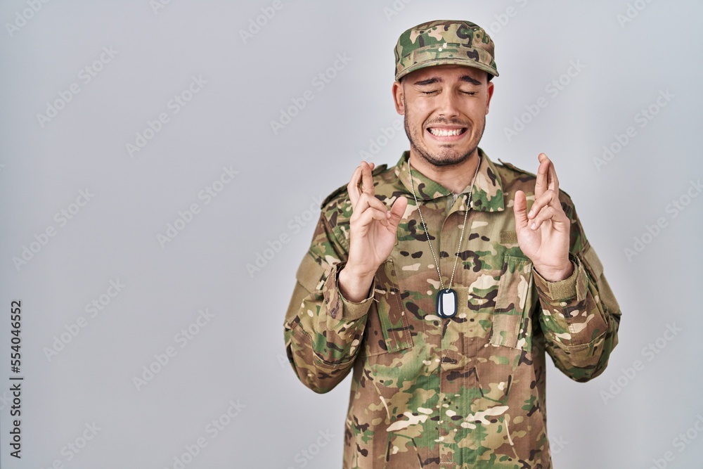 Young hispanic man wearing camouflage army uniform gesturing finger crossed smiling with hope and eyes closed. luck and superstitious concept. Young hispanic man wearing camouflage army uniform gesturing finger crossed smiling with hope and eyes closed. luck and superstitious concept.