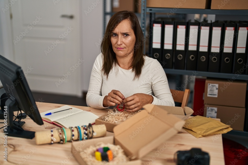 Fototapeta premium Middle age woman working at small bracelets business ecommerce skeptic and nervous, frowning upset because of problem. negative person.