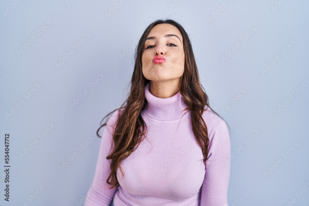 Young brunette woman standing over blue background looking at the camera blowing a kiss on air being lovely and sexy. love expression. Young brunette woman standing over blue background looking at the camera blowing a kiss on air being lovely and sexy. love expression.