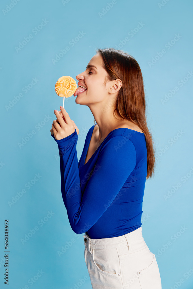 Portrait of beautiful young girl in official stylish clothes posing, eating lollipop over blue background Portrait of beautiful young girl in official stylish clothes posing, eating lollipop over blue background