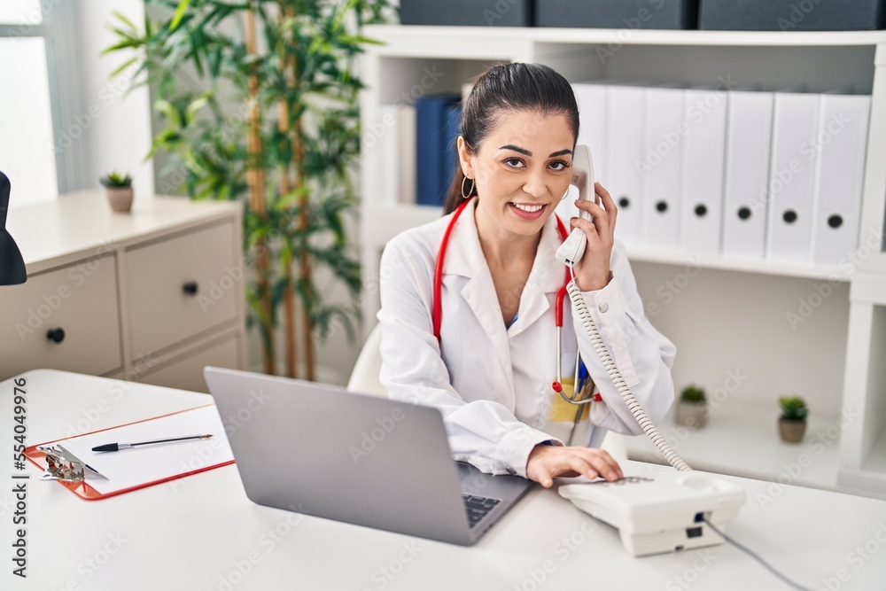 Young beautiful hispanic woman doctor using laptop talking on telephone at clinic Young beautiful hispanic woman doctor using laptop talking on telephone at clinic