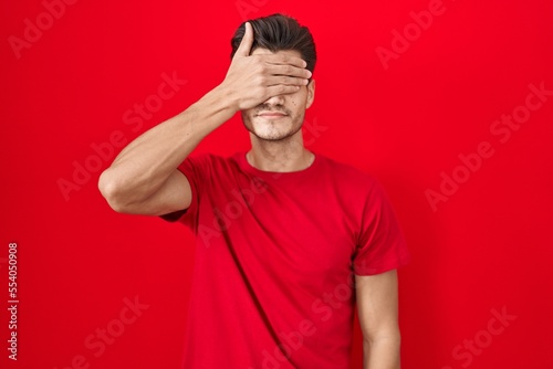 Young hispanic man standing over red background covering eyes with hand, looking serious and sad. sightless, hiding and rejection concept